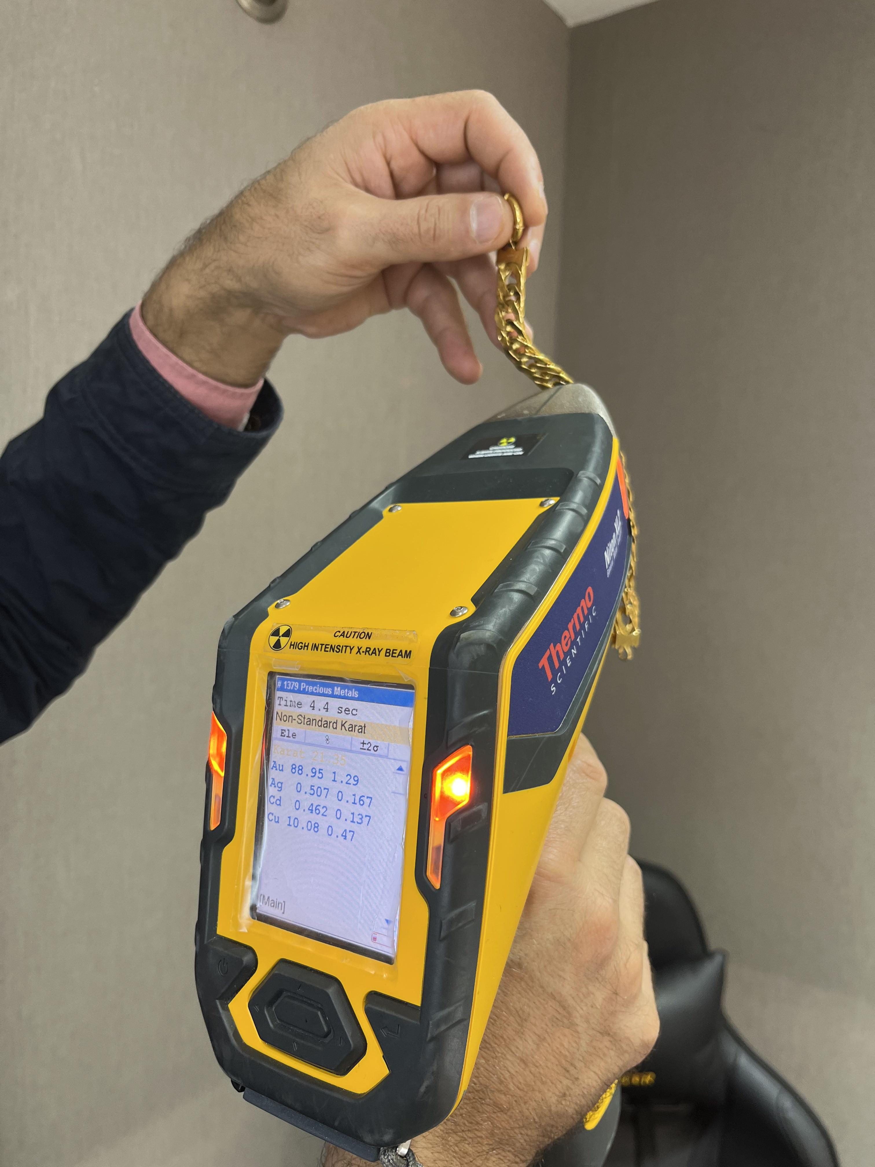 A gold chain held against a handheld XRF analyser, its screen showing the metal content.