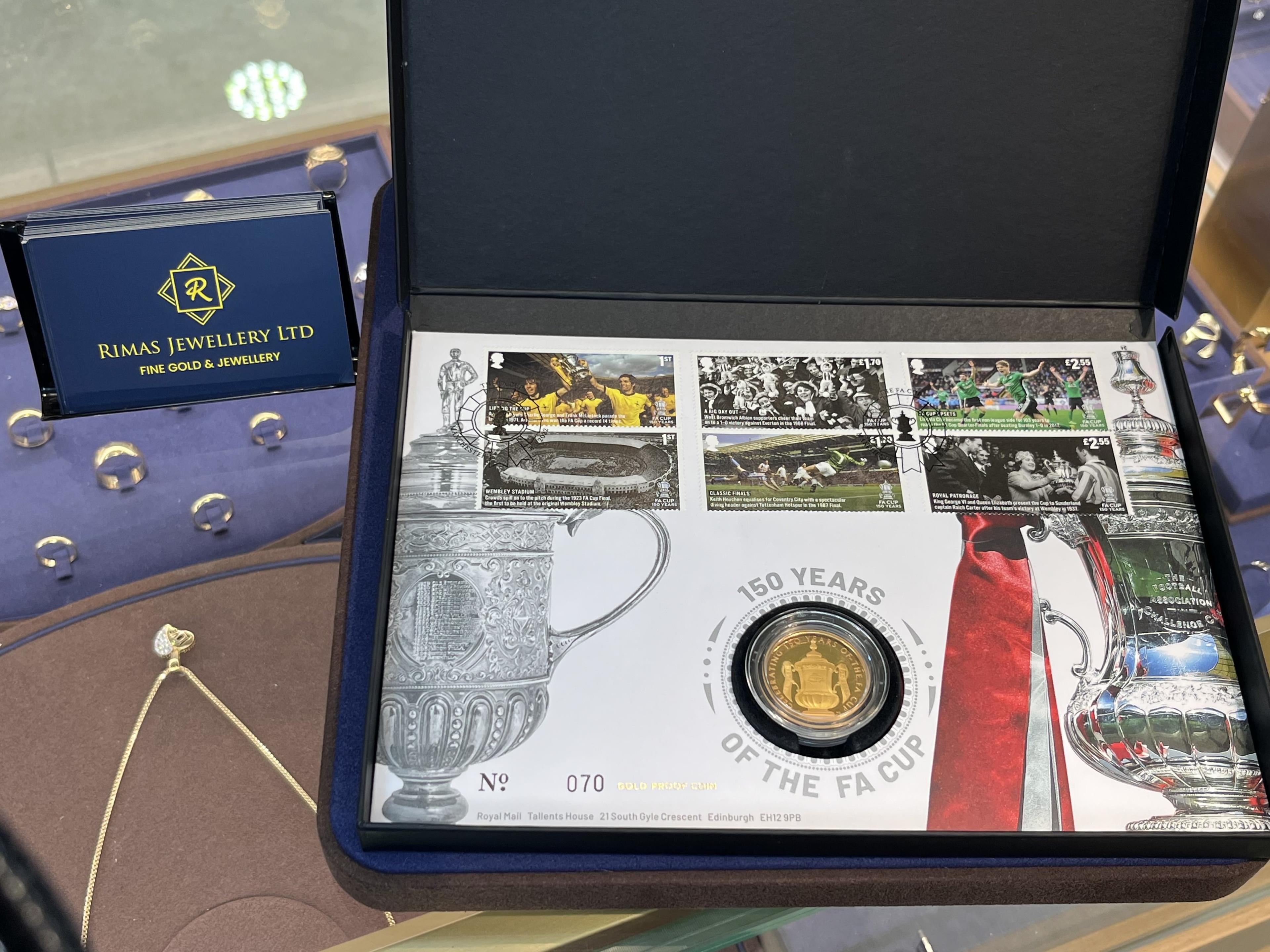 A gold proof coin marking 150 years of the FA Cup, in its numbered presentation cover beside a Rimas Jewellery card.